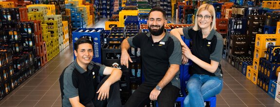 Drei Mitarbeitende sitzen lachend auf gestapelten Getränkekisten in einem Getränkemarkt. Sie tragen schwarze Poloshirts mit grauen Ärmeln und Namensschilder. Der Hintergrund zeigt zahlreiche Getränkekisten in verschiedenen Farben, die ordentlich in Reihen angeordnet sind.