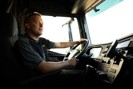 Ein junger Mann sitzt konzentriert hinter dem Steuer eines großen LKWs und schaut nach vorne. Er trägt dunkelgraue Arbeitskleidung und hat eine Tätowierung am rechten Arm. Das moderne Cockpit des Fahrzeugs mit Armaturen und Lenkrad ist ebenfalls gut sichtbar.