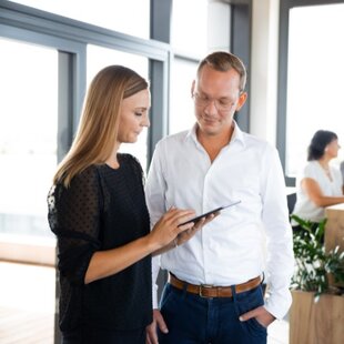 Zwei Mitarbeiter bei einem Meeting in einem modernen Büro. Eine Frau zeigt einem männlichen Kollegen Informationen auf einem Tablet. Im Hintergrund unterhalten sich zwei weitere Personen bei einem Getränk an einem hohen Tisch