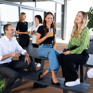 Mehrere Kolleginnen und Kollegen unterhalten sich entspannt auf einer modernen Dachterrasse. Zwei junge Frauen sitzen auf einer Bank und lachen, während sie Getränke in der Hand halten. Ein Mann im weißen Hemd sitzt gegenüber und führt ein Gespräch mit ihnen. Im Hintergrund stehen zwei weitere Frauen mit Gläsern in der Hand und unterhalten sich. Die Atmosphäre wirkt freundlich und offen.