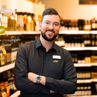 Ein lächelnder Mann in einer dunkelgrauen Uniform steht mit verschränkten Armen vor einem Regal mit Spirituosen und Weinen. Im Hintergrund sind verschiedene Flaschen und Produkte zu sehen. Er trägt eine Armbanduhr und ein Namensschild an seinem Hemd