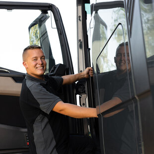 Ein junger Mann in einer dunkelgrauen Arbeitskleidung steht lächelnd an der geöffneten Tür eines LKWs. Er hält sich mit einer Hand an der Tür fest und blickt zur Kamera, während sich sein Gesicht in der glänzenden LKW-Oberfläche spiegelt.