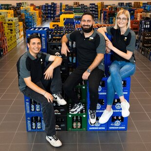 Drei Mitarbeitende sitzen lachend auf gestapelten Getränkekisten in einem Getränkemarkt. Sie tragen schwarze Poloshirts mit grauen Ärmeln und Namensschilder. Der Hintergrund zeigt zahlreiche Getränkekisten in verschiedenen Farben, die ordentlich in Reihen angeordnet sind.