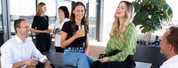 Mehrere Kolleginnen und Kollegen unterhalten sich entspannt auf einer modernen Dachterrasse. Zwei junge Frauen sitzen auf einer Bank und lachen, während sie Getränke in der Hand halten. Ein Mann im weißen Hemd sitzt gegenüber und führt ein Gespräch mit ihnen. Im Hintergrund stehen zwei weitere Frauen mit Gläsern in der Hand und unterhalten sich. Die Atmosphäre wirkt freundlich und offen.