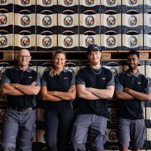 Vier gut gelaunte Mitarbeiterinnen und Mitarbeiter bei einem Fotoshooting vor einer Wand aus Franziskaner Bierkästen, in unserer Unternehmenskleidung.