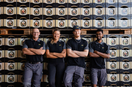Vier gut gelaunte Mitarbeiterinnen und Mitarbeiter bei einem Fotoshooting vor einer Wand aus Franziskaner Bierkästen, in unserer Unternehmenskleidung.