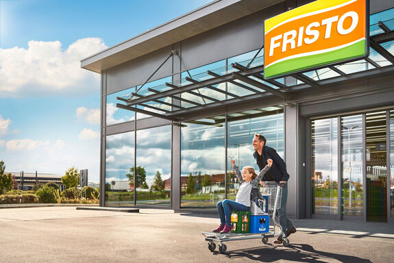 Dieses Bild zeigt den Eingang eines FRISTO Getränkemarktes, wo ein junger Vater mit seiner Tochter nach einem erfolgreichen und tollen Einkaufserlebnis auf einem Einkaufswagen den Markt verlassen.