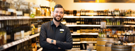 Zu sehen ist einn junger Mitarbeiter mit breitem Lächeln, gut gelaunt, in einem unserer FRISTO Getränkemärkte, im Hintergrund unser umfangreiches Weinsortiment