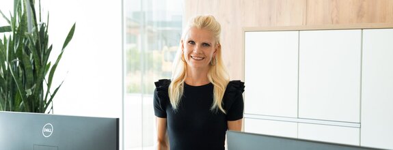 Eine freundliche Frau mit blonden Haaren steht in einem modernen Büro und lächelt in die Kamera. Im Vordergrund sind zwei Computerbildschirme zu sehen, im Hintergrund eine große grüne Pflanze sowie weiße Schränke mit Holzverkleidung. Durch die großen Fenster fällt viel Tageslicht in den Raum, was eine helle und einladende Atmosphäre schafft.